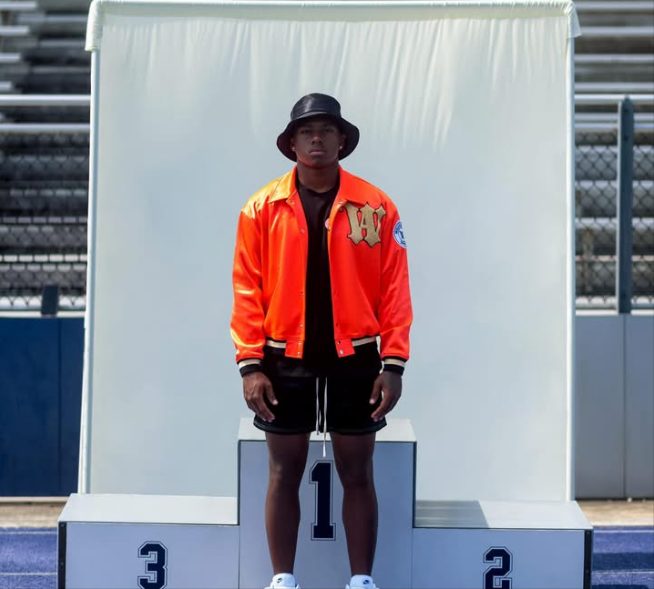 A model on a first place podium in a stadium wearing an orange Willie Austin jacket with a W